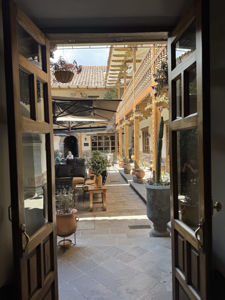View through a rustic wooden doorway into a serene courtyard at Antigua Casona in Cusco, Peru, featuring stone tiles, potted plants, and cozy seating under traditional wooden beams — a tranquil setting that blends Peruvian architecture with warm hospitality.