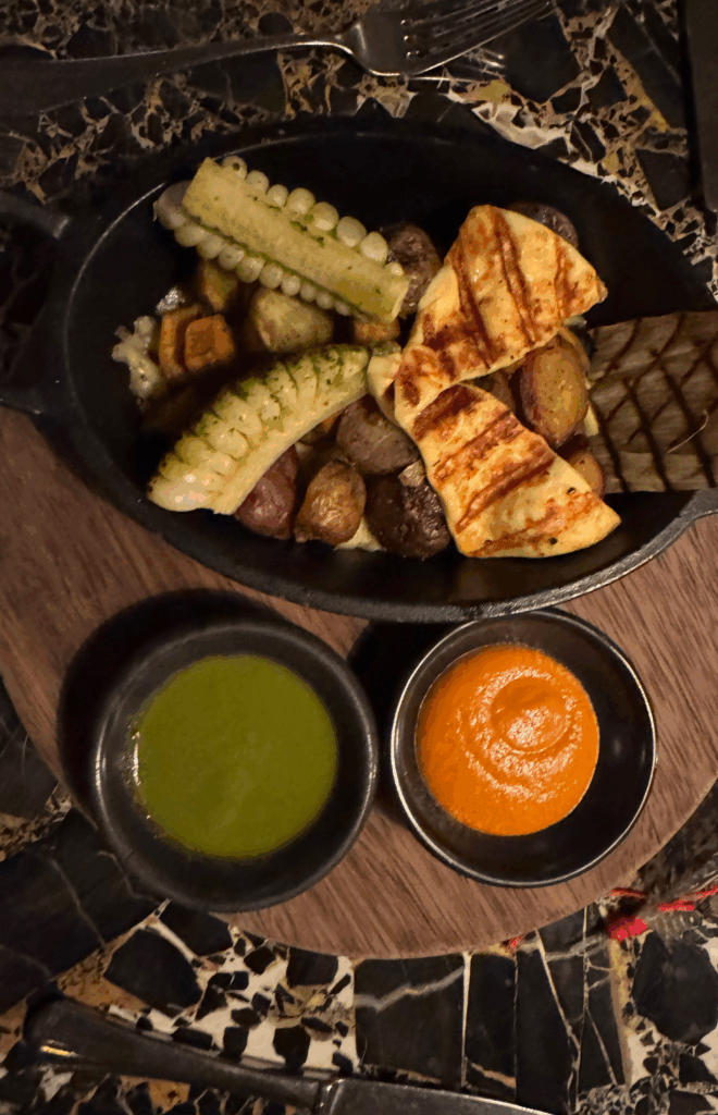 Grilled baby corn, roasted potatoes, and halloumi cheese served with green and red sauces on a rustic wooden board — a vibrant vegetarian dish from Piedra y Sol Restaurant in Cusco, Peru.”