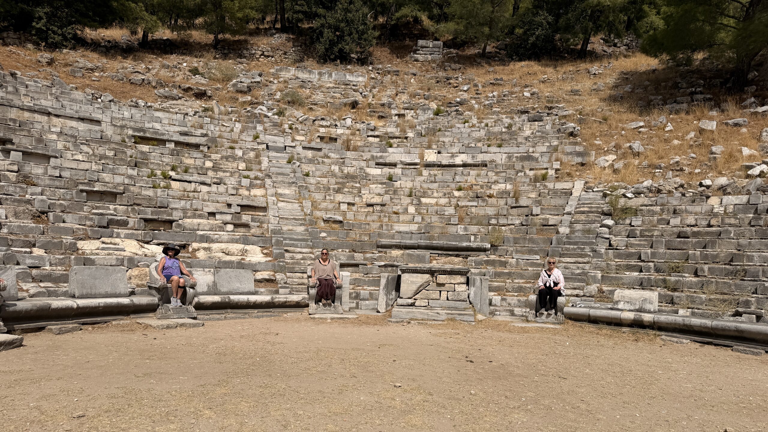 Starseed seated in the front row of the ancient theater of Priene, with tiers of stone seating rising up the hillside behind them.